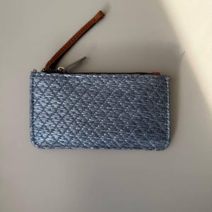 May include: A rectangular, light blue woven wallet with a brown leather zipper pull. The wallet has a silver zipper and a woven texture. The wallet is designed to hold cards and cash.