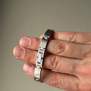 May include: A silver-coloured metal bracelet featuring a rectangular link design. The bracelet is held in a hand, highlighting its polished finish and the way the links articulate. The bracelet is approximately 17.78 cm in length.