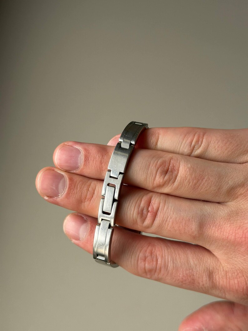 May include: A silver-coloured metal bracelet featuring a rectangular link design. The bracelet is held in a hand, highlighting its polished finish and the way the links articulate. The bracelet is approximately 17.78 cm in length.