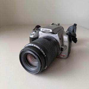 May include: A silver Canon EOS Rebel XT digital SLR camera with a black EF 80-200mm 1:4.5-5.6 II zoom lens attached. The camera body features the Canon logo on the top prism and the Rebel XT model name printed in red and black on the front.