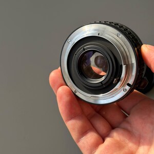 Op de afbeelding: Een close-up van een cameralens met een zilveren en zwarte metalen behuizing. De lens heeft een cirkelvormig ontwerp met een glazen element in het midden. De lens wordt in een hand gehouden.