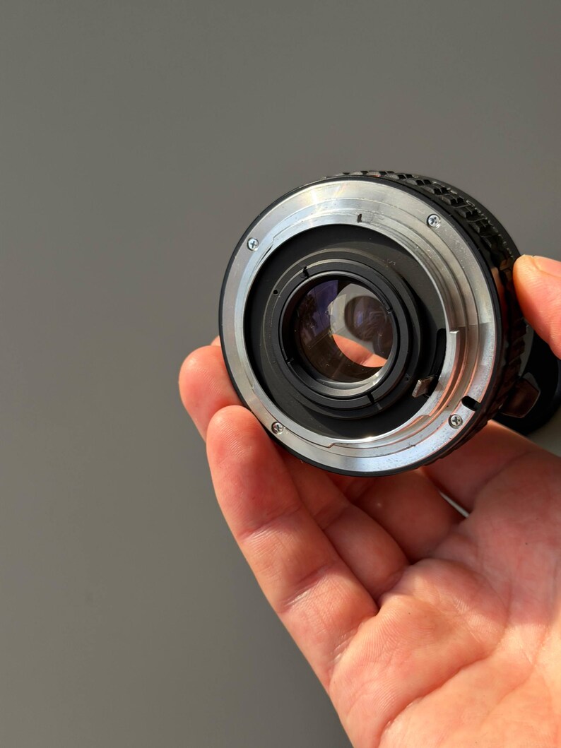 Op de afbeelding: Een close-up van een cameralens met een zilveren en zwarte metalen behuizing. De lens heeft een cirkelvormig ontwerp met een glazen element in het midden. De lens wordt in een hand gehouden.