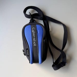 May include: A black and blue Cyber-shot camera case with a black strap. The case has the word "Cyber-shot" printed vertically in white and the brand name "SONY" printed in white. The case is made of a soft material.