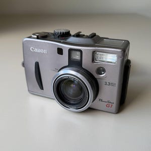 May include: A silver Canon PowerShot G1 digital camera featuring a 3.3 mega pixels lens, a built-in flash, and a Canon zoom lens with 7-21mm 1:2.0-2.5 specifications. The compact camera body includes a top-mounted dial and a shutter button.