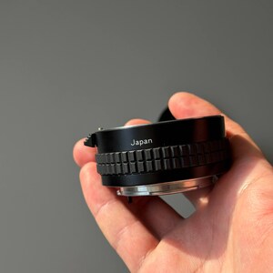 Op de afbeelding: Een zwarte camera lens adapter met een geribbelde focusring. Het woord "Japan" staat op de zijkant van de adapter. De adapter wordt in een hand gehouden tegen een grijze achtergrond.
