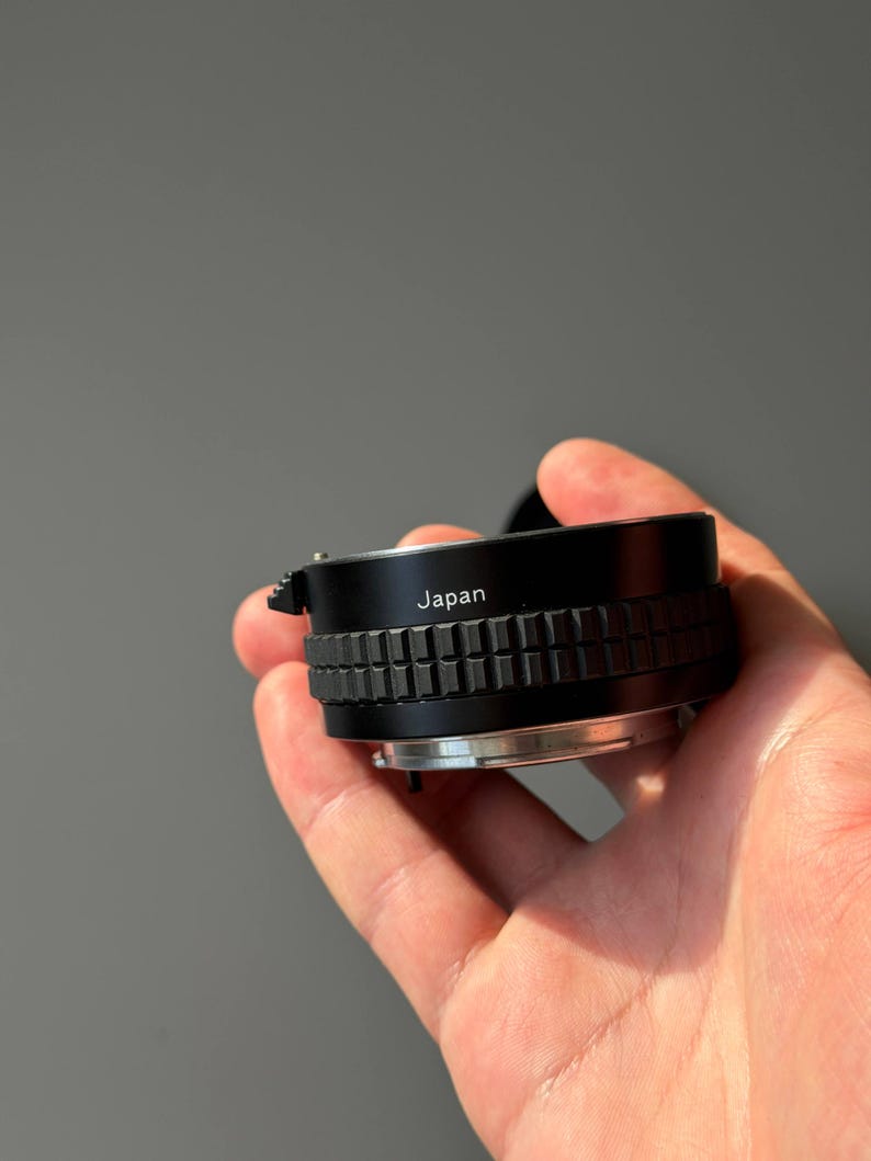 Op de afbeelding: Een zwarte camera lens adapter met een geribbelde focusring. Het woord "Japan" staat op de zijkant van de adapter. De adapter wordt in een hand gehouden tegen een grijze achtergrond.