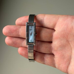 May include: A silver Citizen Elegance wristwatch with a rectangular face and a light blue mother-of-pearl dial. The watch has a silver band and a minimalist design, suitable for everyday wear.