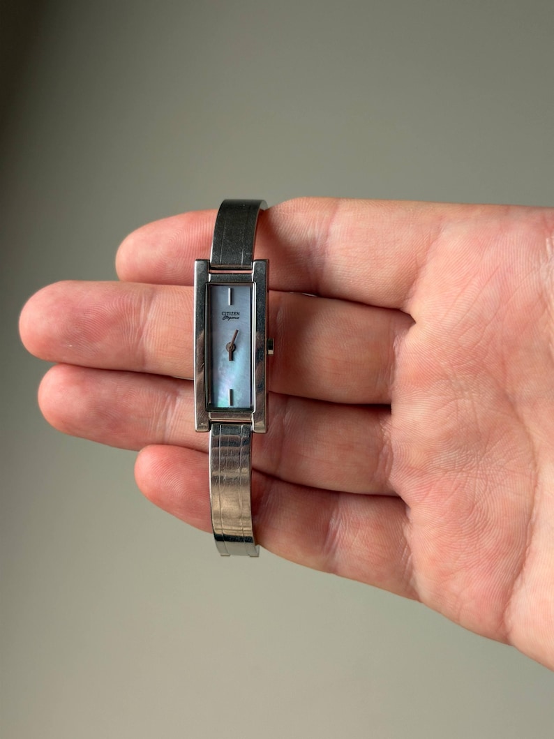 May include: A silver Citizen Elegance wristwatch with a rectangular face and a light blue mother-of-pearl dial. The watch has a silver band and a minimalist design, suitable for everyday wear.