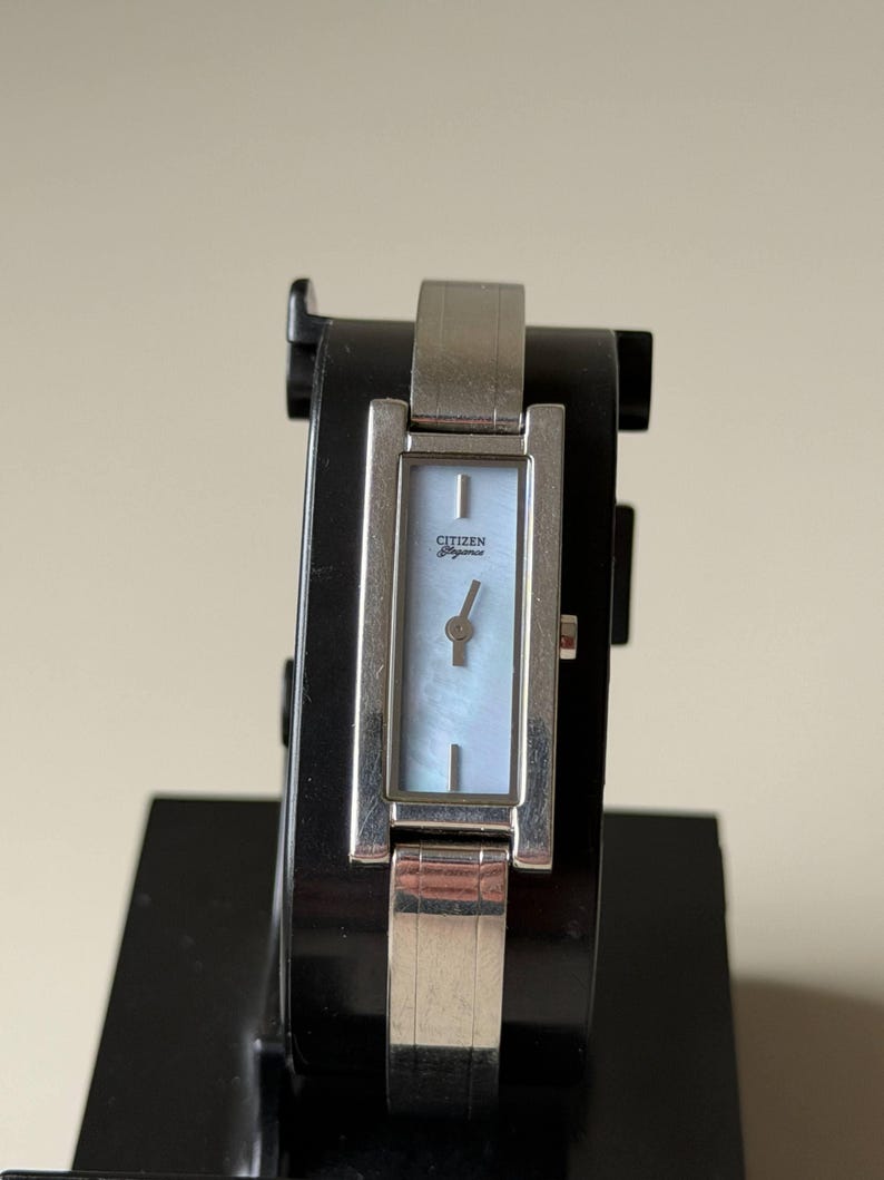 May include: A silver Citizen watch with a rectangular face and a light blue mother-of-pearl dial. The watch has a silver strap and is displayed on a black stand. The watch face has the Citizen logo.