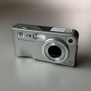 May include: Silver HP Photosmart M415 digital camera. Features a 3X optical zoom lens, 5.2 megapixels, and a built-in flash. The lens has a focal length of 6.0 mm - 18.0 mm. The camera has the HP logo and model name printed on the front.