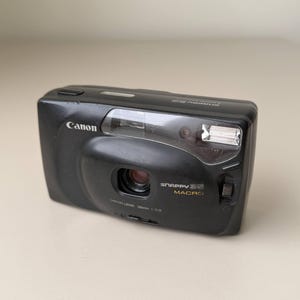May include: A black Canon Snappy 35 Macro point-and-shoot film camera. The front features a central lens, a built-in flash, and the text Canon, Snappy 35 Macro, and Canon Lens 35mm 1:3.8. The camera has a rectangular body with rounded edges.