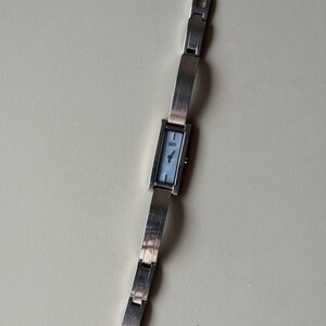 May include: A silver wristwatch with a rectangular face and a light blue dial. The watch has a linked band and a clasp closure. The watch is displayed on a neutral background.