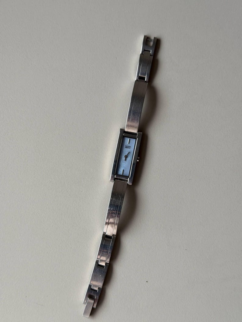 May include: A silver wristwatch with a rectangular face and a light blue dial. The watch has a linked band and a clasp closure. The watch is displayed on a neutral background.