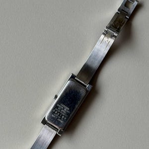 May include: A silver Citizen wristwatch with a rectangular watch face and a stainless steel strap. The watch face has the text "Citizen Water Resist St. Steel 5420-R30223 EBY-11 000109 Mov't Japan". The watch is on a light-coloured surface.