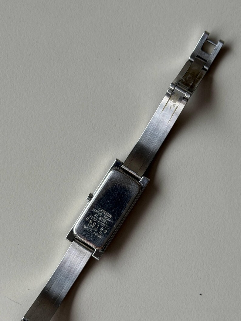 May include: A silver Citizen wristwatch with a rectangular watch face and a stainless steel strap. The watch face has the text "Citizen Water Resist St. Steel 5420-R30223 EBY-11 000109 Mov't Japan". The watch is on a light-coloured surface.