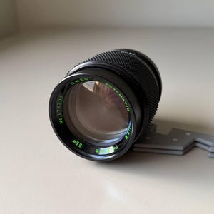 May include: A black camera lens with green text, including the brand name "OWEN" and the specifications "55mm" and "f=1:3.5m". The lens has a textured grip and is mounted on a gray support.
