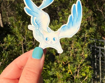 May include: A die-cut sticker featuring a light-coloured, winged feline creature with large blue eyes and blue-tipped wings. The illustration has a soft, watercolour-like appearance with a white border, depicting the creature in a flying pose.