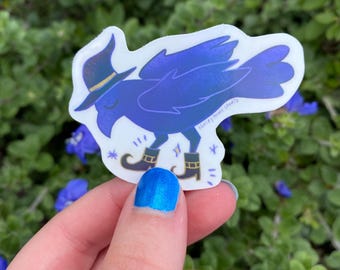 May include: A die-cut sticker featuring a dark blue crow wearing a pointed hat and curled-toe boots. The crow has closed eyes and is surrounded by small star accents. The text Haley Howe Studio is printed in small letters on the wing.