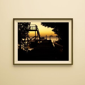 May include: A framed photograph of a pier at sunset. The pier extends into the water, with a small structure at the end. The sky is a gradient of yellow and orange. Silhouetted trees frame the scene.