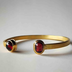 Puede incluir: Una pulsera de brazalete abierto en tono dorado con dos piedras preciosas de granate rojo oscuro, pulidas y de forma ovalada, engastadas en cada extremo. La banda de metal tiene un acabado liso y mate con un diseño curvo y abierto.