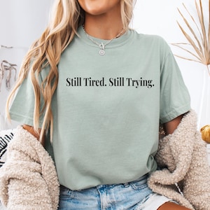 May include: A light green t-shirt with the text "Still Tired. Still Trying." in black. The shirt is paired with denim shorts and a cream-colored cardigan. The model is wearing a silver necklace.