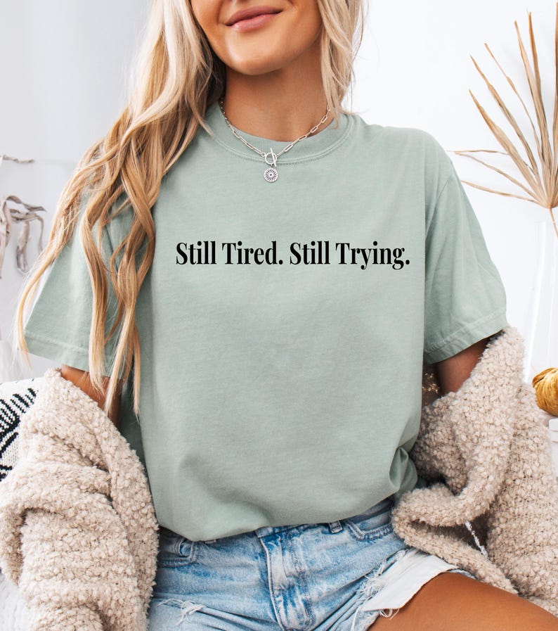 May include: A light green t-shirt with the text "Still Tired. Still Trying." in black. The shirt is paired with denim shorts and a cream-colored cardigan. The model is wearing a silver necklace.