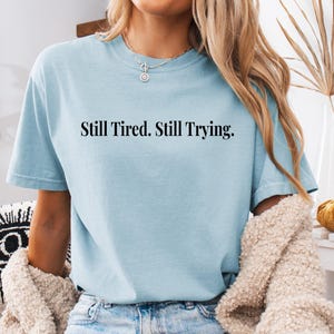 May include: A light blue t-shirt with the text "Still Tired. Still Trying." in black. The shirt is paired with blue jeans and a cream-colored sweater. The overall aesthetic is casual and comfortable.