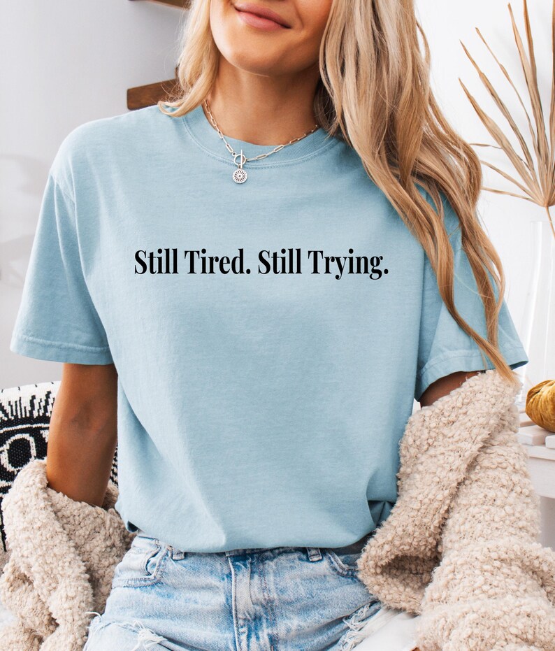 May include: A light blue t-shirt with the text "Still Tired. Still Trying." in black. The shirt is paired with blue jeans and a cream-colored sweater. The overall aesthetic is casual and comfortable.