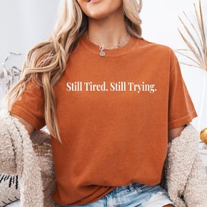 May include: Burnt orange t-shirt with the text "Still Tired. Still Trying." The shirt has short sleeves and a crew neck. The shirt is made of a soft, comfortable material.