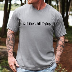 May include: A gray t-shirt with the text "Still Tired. Still Trying." in black. The shirt is a solid color and has a crew neck. The person wearing the shirt has tattoos on their arms.
