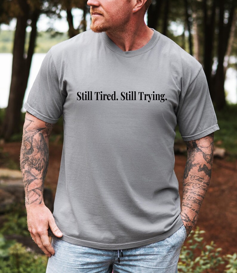 May include: A gray t-shirt with the text "Still Tired. Still Trying." in black. The shirt is a solid color and has a crew neck. The person wearing the shirt has tattoos on their arms.