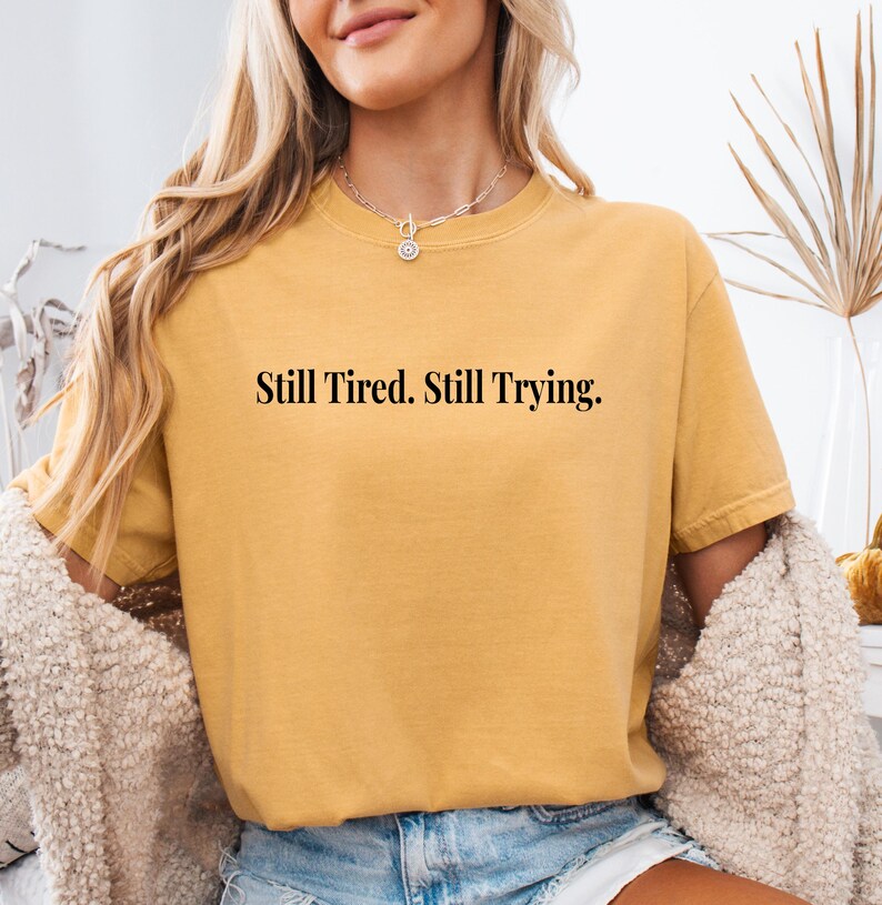May include: Mustard yellow t-shirt with the text "Still Tired. Still Trying." in a black, simple font. The shirt has short sleeves and a crew neck. A woman is wearing the shirt, with a cream-colored sweater draped over her shoulders.