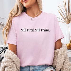 May include: A light pink t-shirt with the text "Still Tired. Still Trying." in black. The shirt has short sleeves and a crew neck. The model is wearing a necklace and a beige sweater.