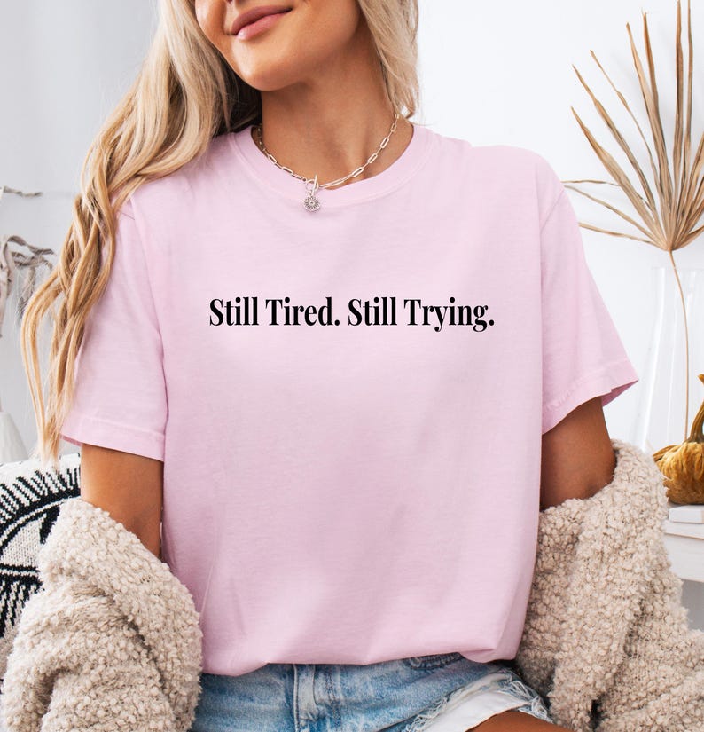 May include: A light pink t-shirt with the text "Still Tired. Still Trying." in black. The shirt has short sleeves and a crew neck. The model is wearing a necklace and a beige sweater.