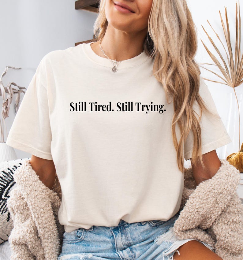 May include: Cream-colored t-shirt with the text "Still Tired. Still Trying." in a black, bold font. The shirt is a relaxed fit and appears to be made of a soft material. The model is wearing a cream-colored sweater.