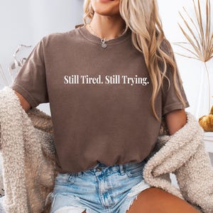 May include: A brown t-shirt with the text "Still Tired. Still Trying." in white. The shirt is paired with denim shorts and a cream-colored cardigan. The model is wearing a silver necklace.