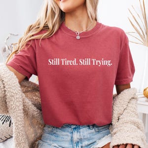 May include: A maroon t-shirt with the text "Still Tired. Still Trying." in white. The shirt has short sleeves and a crew neck. The person is wearing blue denim shorts and a cream-colored cardigan.