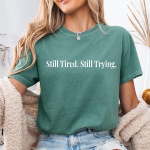 May include: A teal-colored t-shirt with the text "Still Tired. Still Trying." in white. The shirt has short sleeves and a crew neck. The model is wearing a cream-colored cardigan and blue jeans.