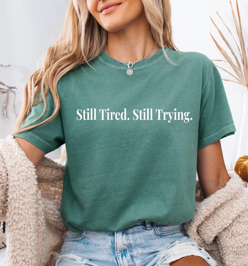 May include: A teal-colored t-shirt with the text "Still Tired. Still Trying." in white. The shirt has short sleeves and a crew neck. The model is wearing a cream-colored cardigan and blue jeans.