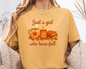 May include: Mustard yellow t-shirt with the text "Just a girl who loves fall." The graphic includes a coffee mug with a leaf, a candle, and books. The shirt is a comfortable, casual style.