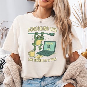 May include: Cream-colored t-shirt with the text "Debugging Life" and a cartoon frog hanging from a cable. A laptop displays an error message. A coffee cup and a "Fix Later" note are also visible.