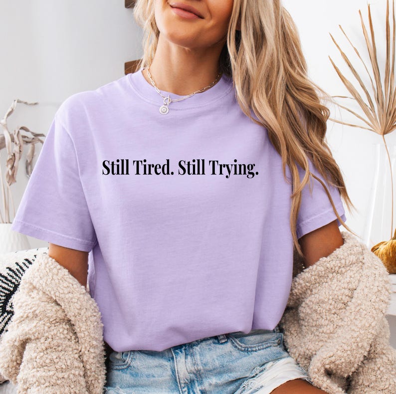May include: A light purple t-shirt with the text "Still Tired. Still Trying." in black. The shirt is a crew neck style and appears to be made of a soft material. The shirt is worn with denim shorts and a cream-colored cardigan.