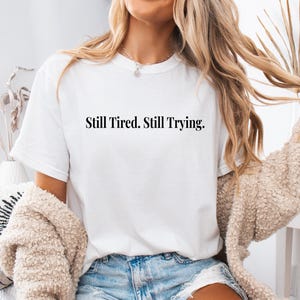 May include: White t-shirt with the text "Still Tired. Still Trying." in a black, simple font. The shirt is worn by a person with long blonde hair and a beige sweater.