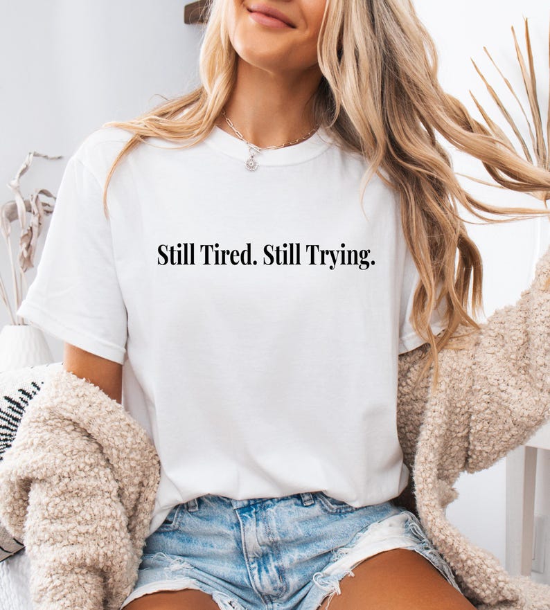 May include: White t-shirt with the text "Still Tired. Still Trying." in a black, simple font. The shirt is worn by a person with long blonde hair and a beige sweater.