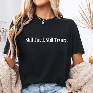 May include: Black t-shirt with the white text "Still Tired. Still Trying." The shirt is a crew neck style. The model is wearing a silver necklace and a beige cardigan.