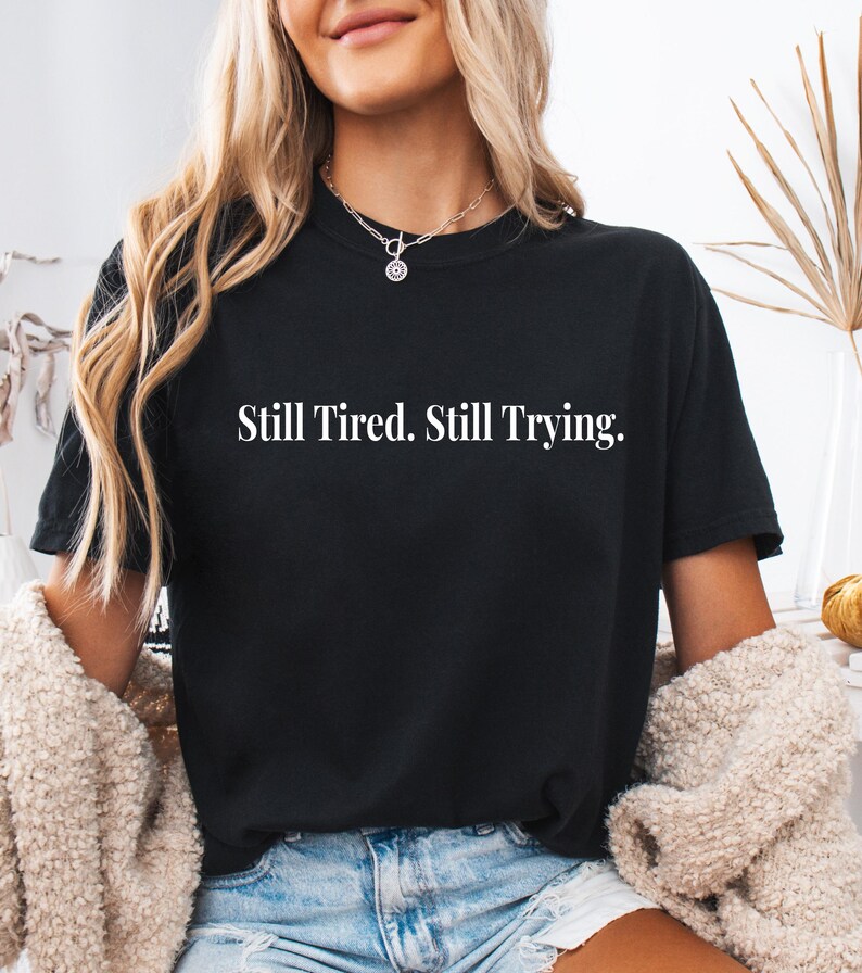 May include: Black t-shirt with the white text "Still Tired. Still Trying." The shirt is a crew neck style. The model is wearing a silver necklace and a beige cardigan.