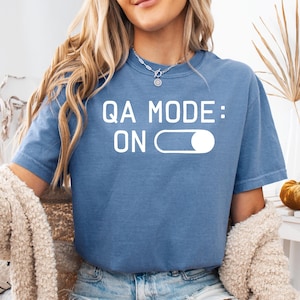May include: A blue t-shirt with the text "QA MODE: ON" in white. The design includes a white on/off slider graphic. The shirt is a solid color and appears to be made of a soft material.