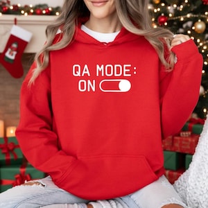 May include: A red hoodie with the text "QA MODE: ON" in white. The hoodie has a white drawstring and a front pocket. The background includes Christmas decorations, such as a stocking and a lit Christmas tree.