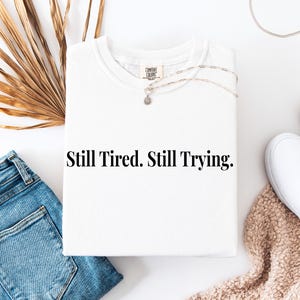 May include: A white t-shirt with the text "Still Tired. Still Trying." is displayed flat on a white surface. The shirt is paired with blue jeans, white sneakers, and a necklace. A dried palm leaf and a beige blanket are also in the image.