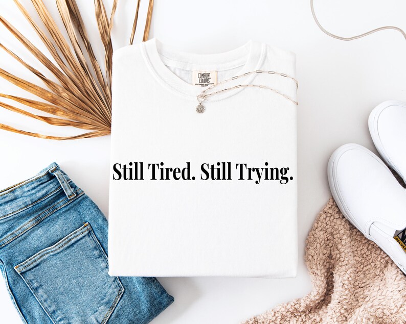 May include: A white t-shirt with the text "Still Tired. Still Trying." is displayed flat on a white surface. The shirt is paired with blue jeans, white sneakers, and a necklace. A dried palm leaf and a beige blanket are also in the image.